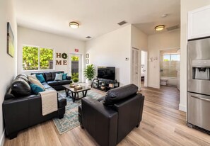 A modern living room with a sectional sofa, a flat-screen TV, and a kitchen area with a stainless steel refrigerator.