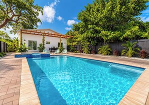 A swimming pool with a wooden deck, surrounded by lush greenery and a white building with a pergola.