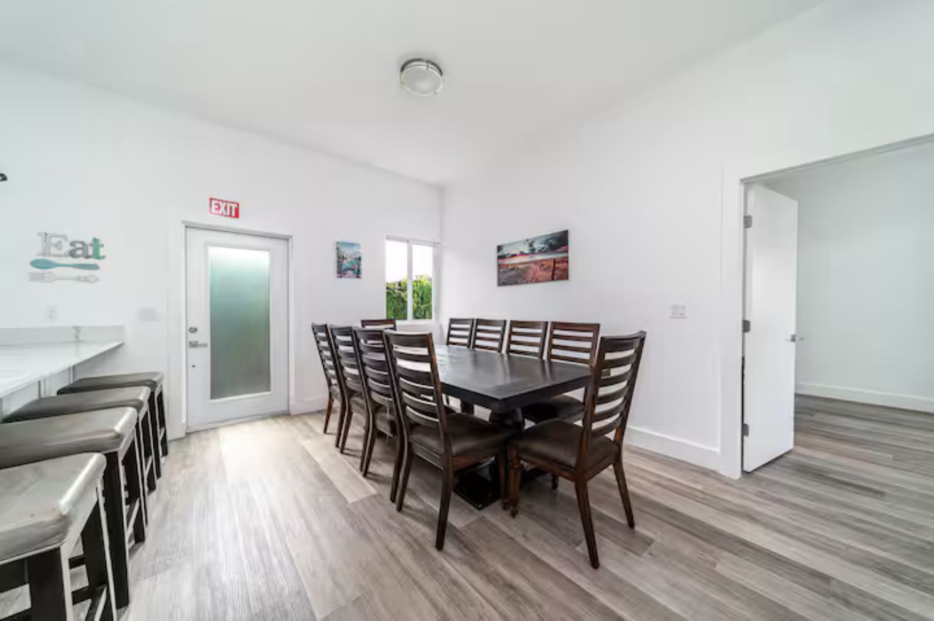 A modern dining area with a large table and chairs, a white door with an exit sign, and a view of greenery outside.