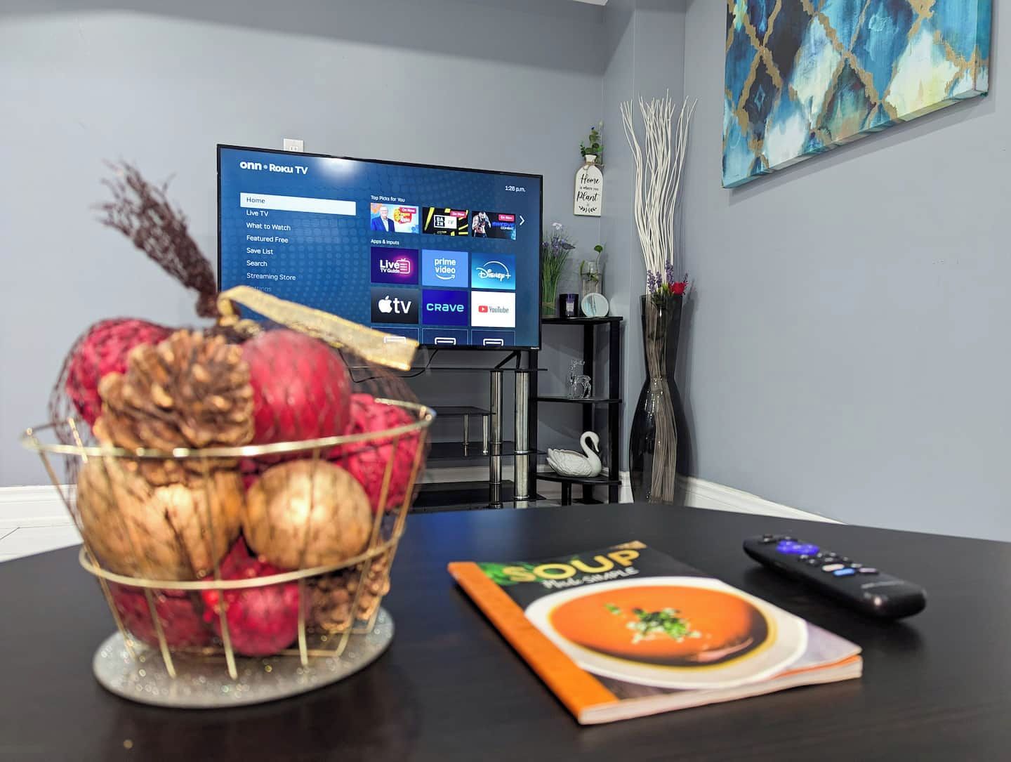 Een kamer met een televisie waarop een streamingdienst draait, een schaal fruit, een afstandsbediening en een boek op de tafel.