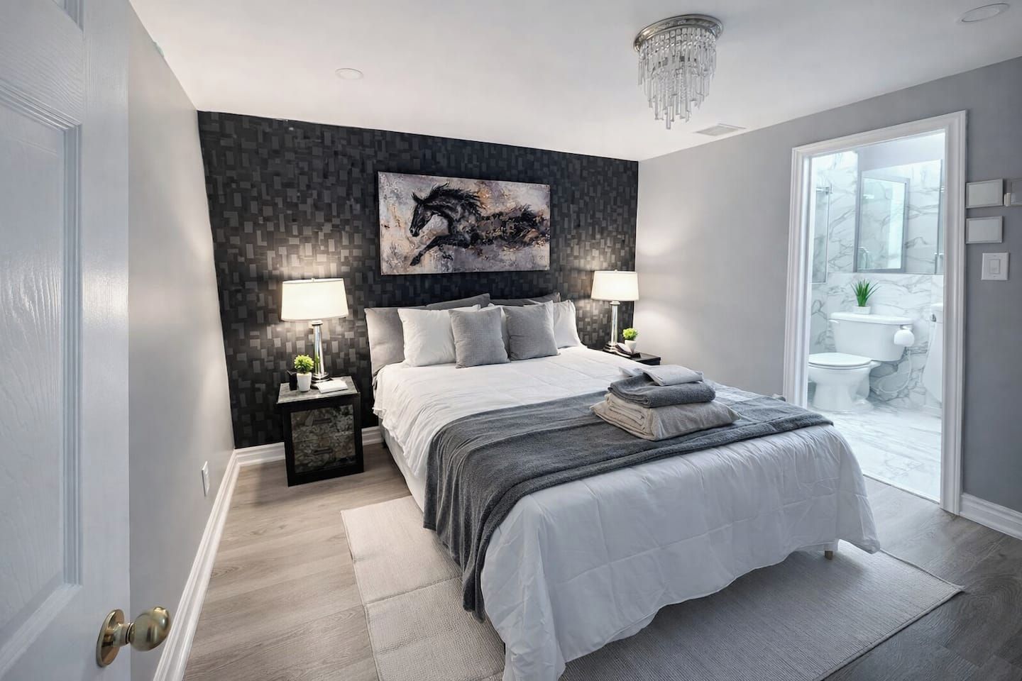 A modern bedroom with a large bed, two bedside lamps, a chandelier, and a bathroom visible through an open door.