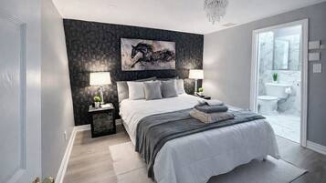 A modern bedroom with a large bed, two bedside lamps, a chandelier, and a bathroom visible through an open door.