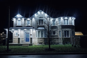 Une maison en pierre à deux étages, avec une porte bleue et de nombreuses fenêtres, éclairée par des lampes extérieures.