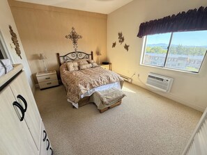 A bedroom with a bed, bedside tables, a dresser, a window with a view, and a wall-mounted air conditioner. - Stunning Lake Winnipesaukee Views, 2BR + Pool, Walk to Weirs Beach (Laconia)