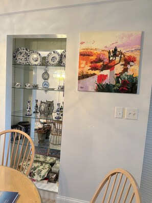 A dining area with a glass-top table, wooden chairs, and a wall-mounted painting. - Stunning Lake Winnipesaukee Views, 2BR + Pool, Walk to Weirs Beach (Laconia)
