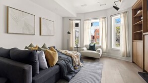 Una sala moderna con un sofá oscuro, un sillón blanco y una alfombra estampada. Hay dos cuadros enmarcados en la pared, una estantería y una ventana con cortinas.