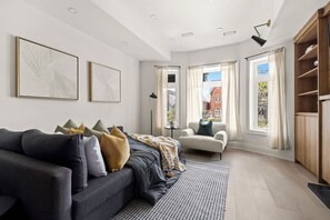 A modern living room with a dark sofa, a white armchair, and a patterned rug. There are two framed artworks on the wall, a bookshelf, and a window with curtains. - Bright West End Gem, Walk to Waterfront! (Toronto)