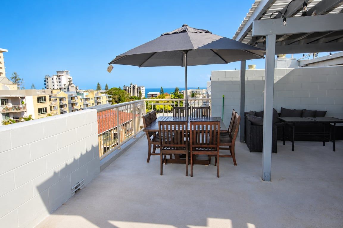 Un balcon avec une table et des chaises, un parasol, et une vue sur la ville et l’océan.