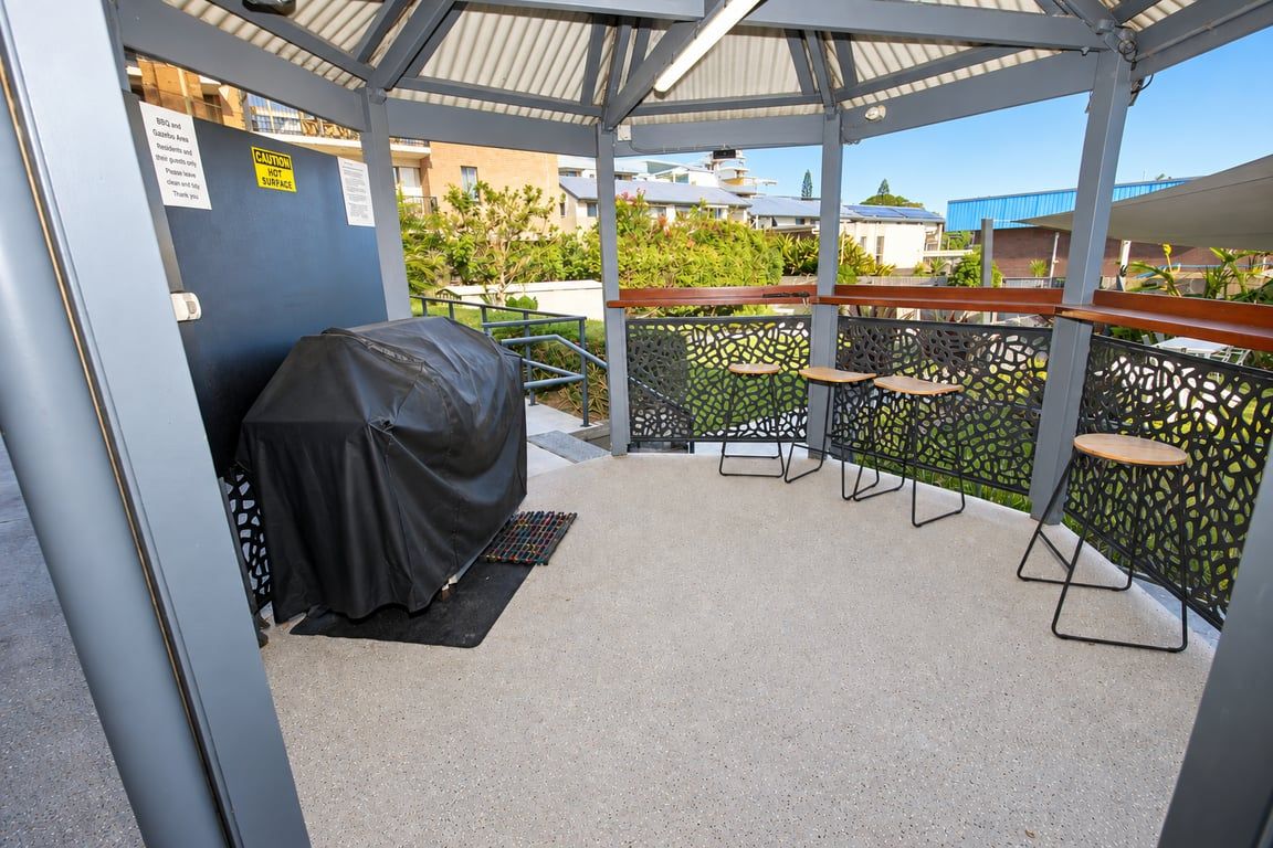 A covered outdoor seating area with tables and chairs, a barbecue grill, and a metal fence.