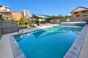 A rectangular swimming pool with a clear blue surface, surrounded by lounge chairs and a wooden fence.