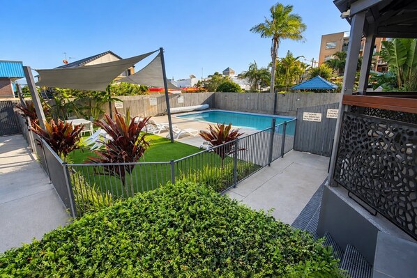 A swimming pool area with a fence, palm trees, and a shaded seating area.