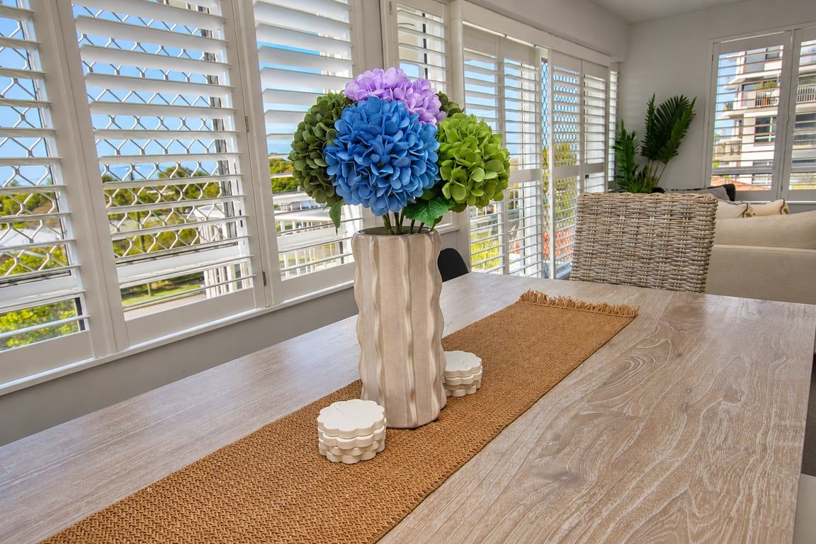 A modern interior with a wooden dining table, a vase with flowers, and a view through large windows.