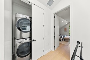 Ein Waschraum mit zwei übereinander gestapelten Waschmaschinen und einer Tür zu einem Schlafzimmer mit einem Bett und einem Fenster.