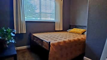 A bedroom with a bed, a window with blinds, a plant, and a chair.