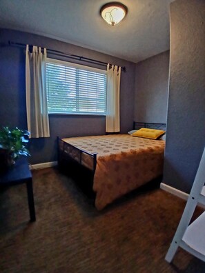 A bedroom with a bed, a window with blinds, a plant, and a chair.
