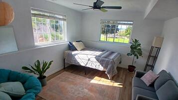 Una habitación con una cama, dos ventanas, un ventilador de techo, una silla azul, un sofá gris y una planta en maceta.