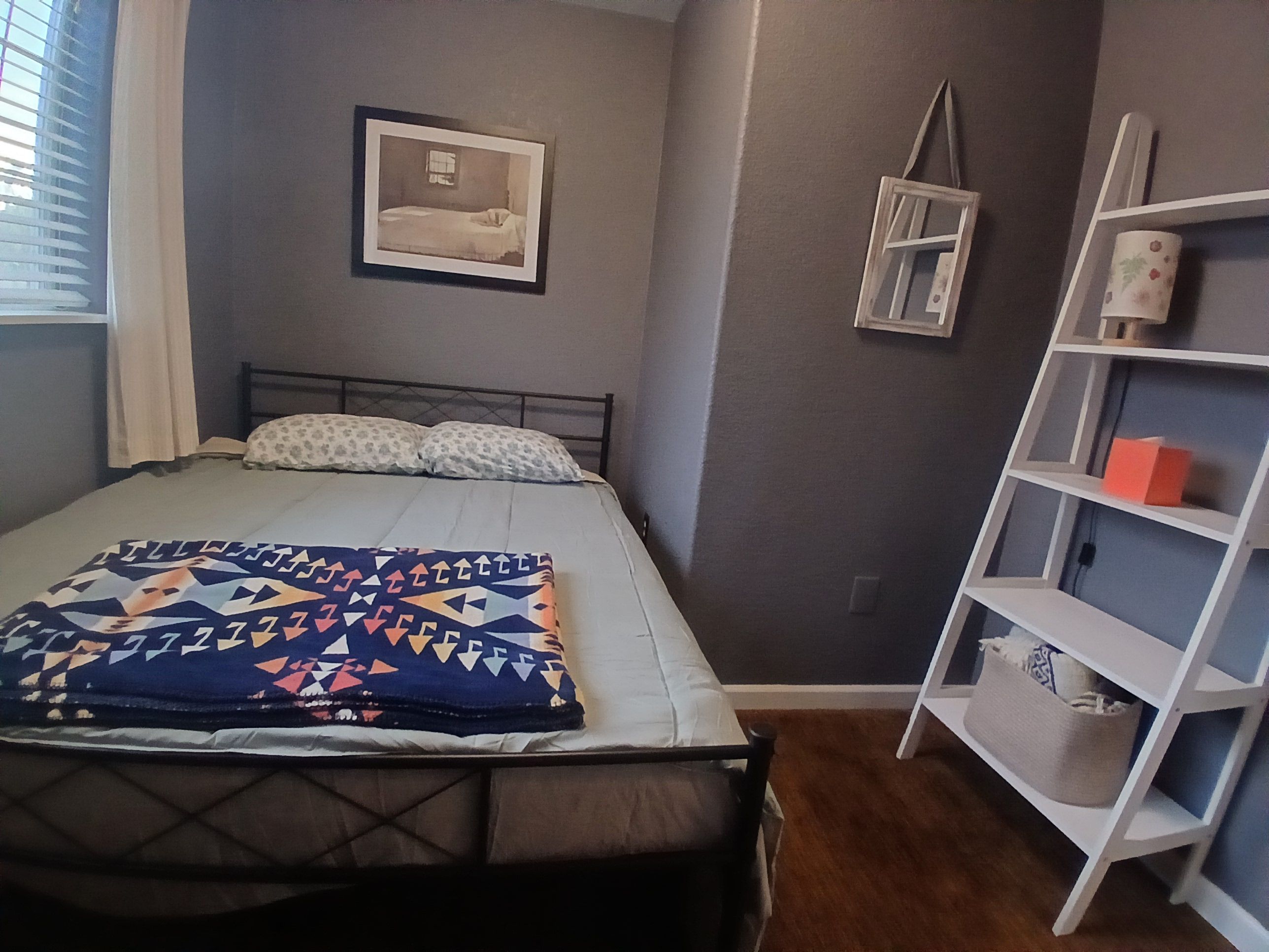 Une chambre à coucher avec un lit, une étagère de chevet, un tableau encadré au mur et une fenêtre avec des rideaux.