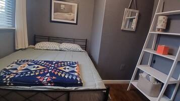 Une chambre à coucher avec un lit, une étagère de chevet, un tableau encadré au mur et une fenêtre avec des rideaux.