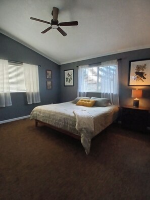 A bedroom with a bed, a ceiling fan, two windows with curtains, and a nightstand with a lamp.