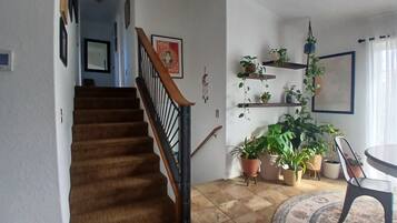 Een trap met houten leuning die uitkomt op een plateau, een eetruimte met een ronde tafel en stoelen, en meerdere potplanten op planken.