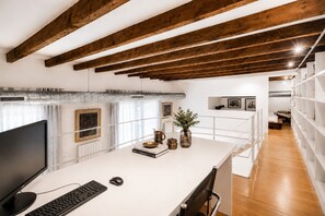 A modern office space with a white desk, computer, and chair. There are books, a vase with plants, and a shelf with framed pictures. The ceiling has exposed wooden beams, and there's a white railing overlooking a bedroom area. - 3 Bedroom, 3 bathrooms Quiet Flat + Cable Internet (Milano)