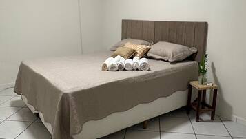 Een slaapkamer met een bed, kussens, een nachtkastje met een plant en een airconditioner aan de muur.