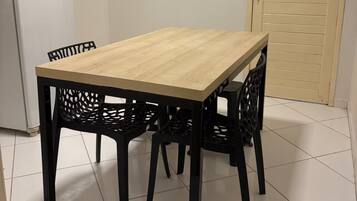 Een eethoek met een houten tafel en zwarte stoelen, een witte koelkast, en een deur voorzien van horizontale jaloezieën.