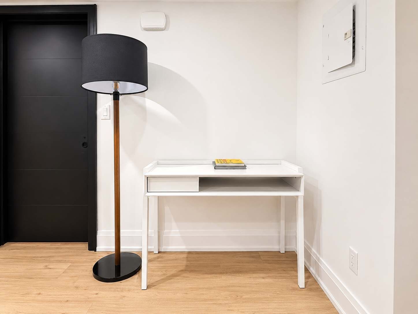 Una habitación con un escritorio blanco, una lámpara de pie negra y una puerta oscura.