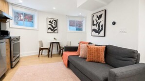 A modern living room with a grey sofa, a small dining table, and a kitchen area with an oven. - Stylish 2BR West End Oasis | Huge Deck near High Park (Toronto)