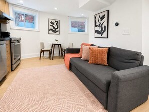 A modern living room with a grey sofa, a small dining table, and a kitchen area with an oven. - Stylish 2BR West End Oasis | Huge Deck near High Park (Toronto)