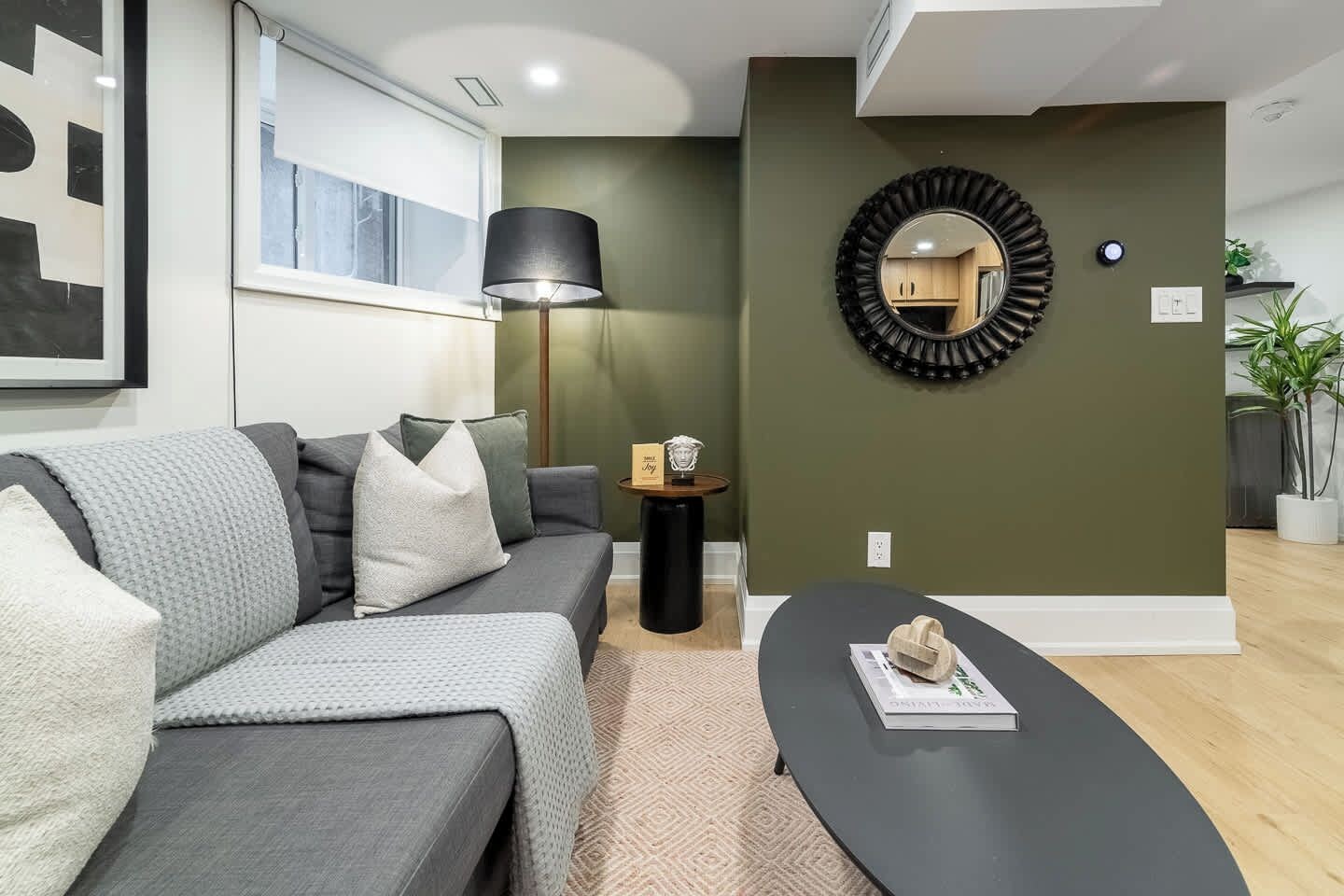A modern living room with a grey sofa, a round mirror, a small table with a lamp, and a potted plant.