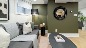 A modern living room with a grey sofa, a round mirror, a small table with a lamp, and a potted plant.