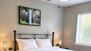 En pænt redt seng med et sengegærde, to natborde med lamper, et vægmonteret tv og et vindue med persienner.