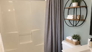 Et badeværelse med badekar, toilet og håndvask. Der er en bruseforhæng, en hylde med håndklæder og en plante, samt en kurv på toilettet.