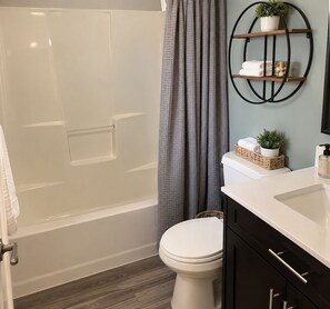 A bathroom with a bathtub, toilet, and a sink. There is a shower curtain, a shelf with towels and a plant, and a basket on the toilet. - Spacious 4-bedroom house in gorgeous Kennewick with AC (Kennewick)