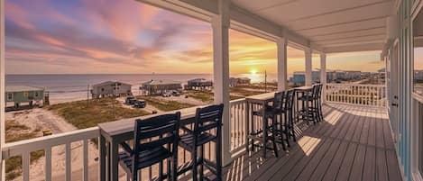 A wooden deck with chairs and a table, overlooking a beach with buildings in the distance.