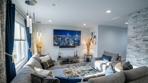 A modern living room with a large sofa set, a flat-screen TV, and a cityscape painting. - Riverfront Front | Fire Pit | Arcade | Fishing| (New Brunswick)