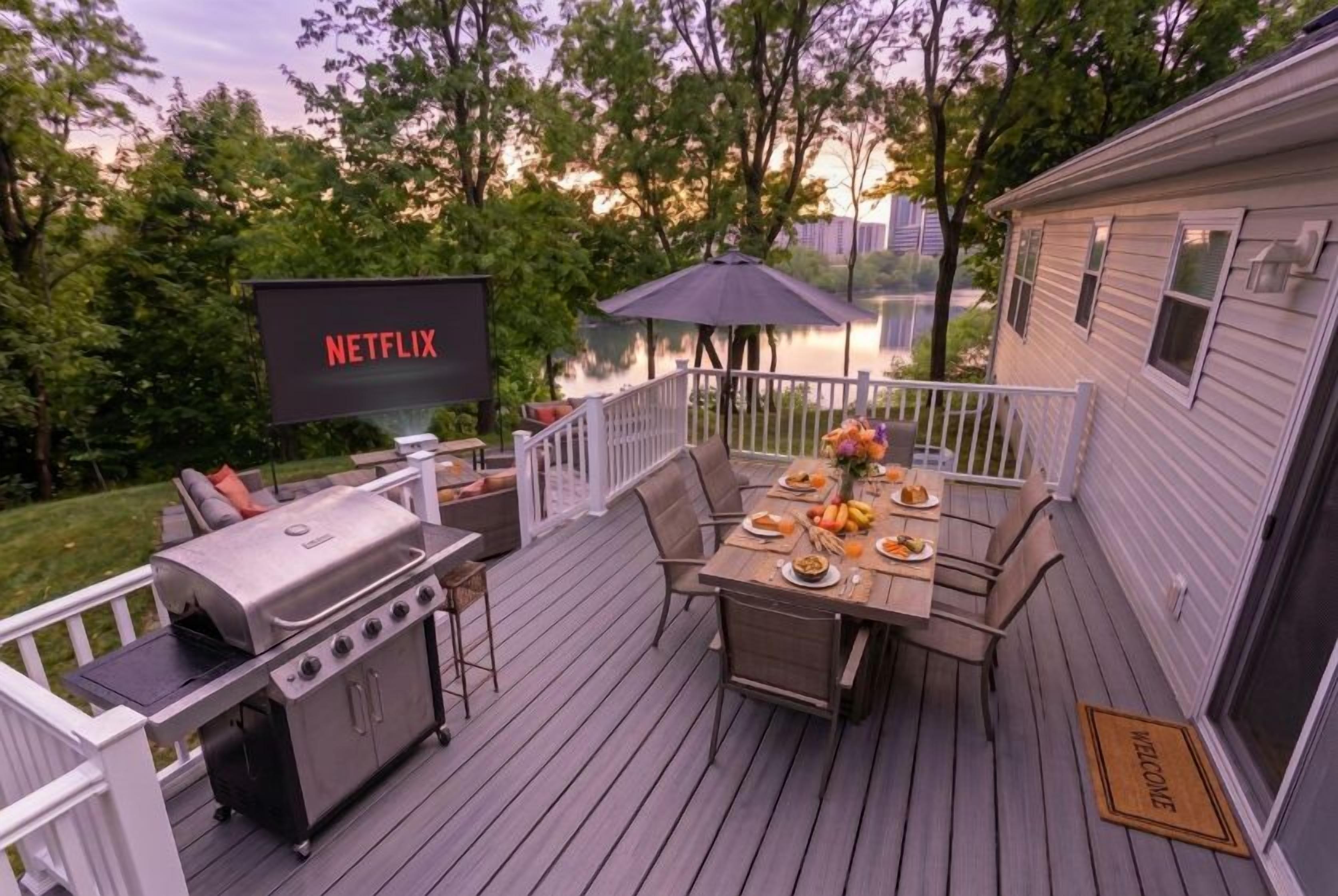 Una terraza con una parrilla, mesa de comedor y sillas, una pantalla de televisión mostrando 'NETFLIX', y vistas a un lago.