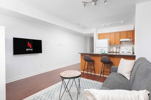 A modern living room with a flat-screen TV mounted on the wall, a grey sofa, two bar stools, and a small round table. - Modern Yonge-Dundas Studio | Steps to Eaton Centre | Parking Available (Toronto)