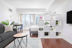 Ein modernes Wohnzimmer mit einer Couch, einem Schreibtisch und einem Sessel. Ein großes Fenster mit Vorhängen, ein an der Wand montierter Fernseher und ein Bücherregal mit Pflanzen sind ebenfalls vorhanden.