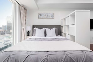 A hotel room with a bed, a nightstand, a lamp, a painting, and a view of the city. - Modern Yonge-Dundas Studio | Steps to Eaton Centre | Parking Available (Toronto)