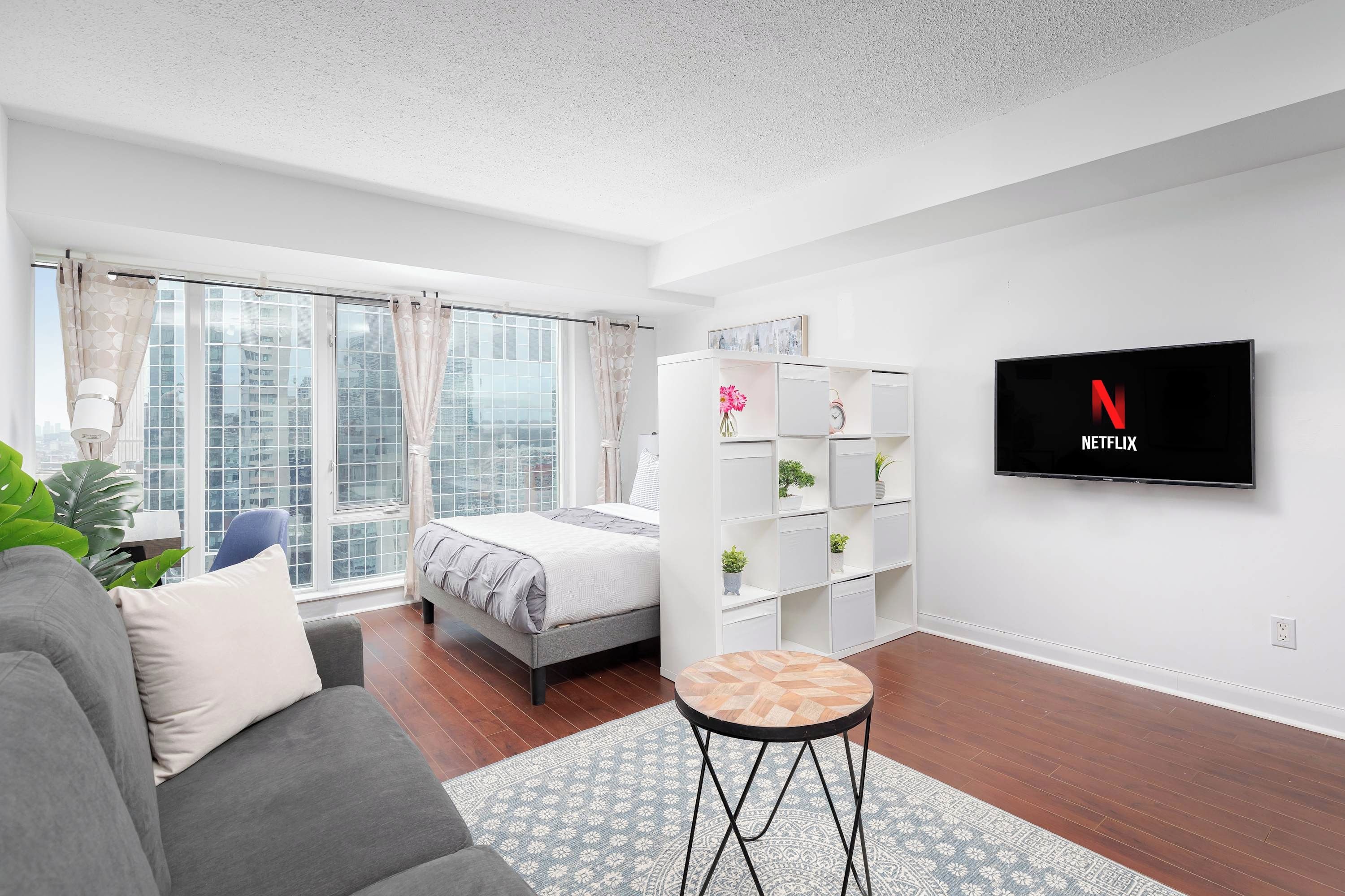 A modern living room with a grey sofa, a round coffee table, a bed with a grey duvet, a TV mounted on the wall, and a window with curtains.