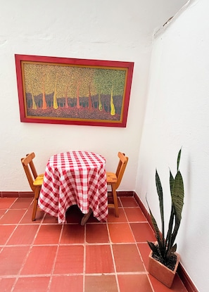 Un pequeño patio con una mesa y sillas a cuadros, una planta en maceta y una obra de arte enmarcada en la pared.