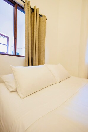 Cama com roupa de cama branca em quarto com janela e cortinas.