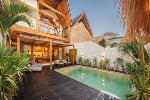 Una casa a due piani in legno con piscina, area salotto esterna e piante tropicali.