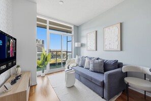 A modern living room with a grey sofa, a white side table, a flat-screen TV, and a large window offering a city view.