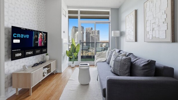 Una sala de estar moderna con un televisor de pantalla plana, un sofá gris, una mesa de centro blanca y un balcón con vistas a la ciudad.