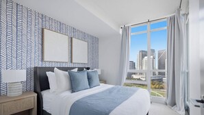 Un dormitorio moderno con una cama grande, una mesita de noche, un televisor montado en la pared y vistas a un paisaje urbano a través de un ventanal amplio.