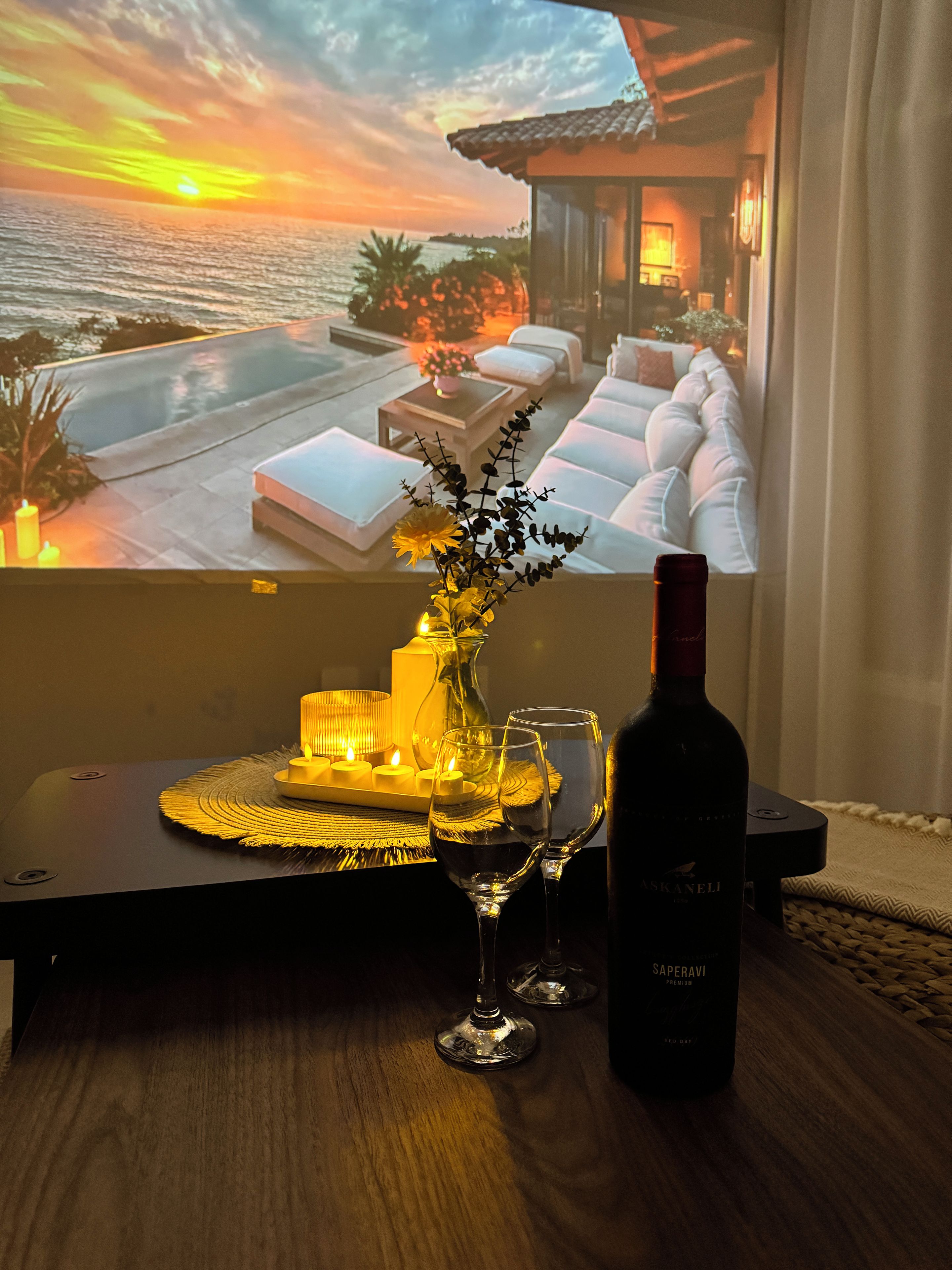 A cozy room with a view of a sunset over the ocean, a table with candles, a bottle of wine, and glasses.