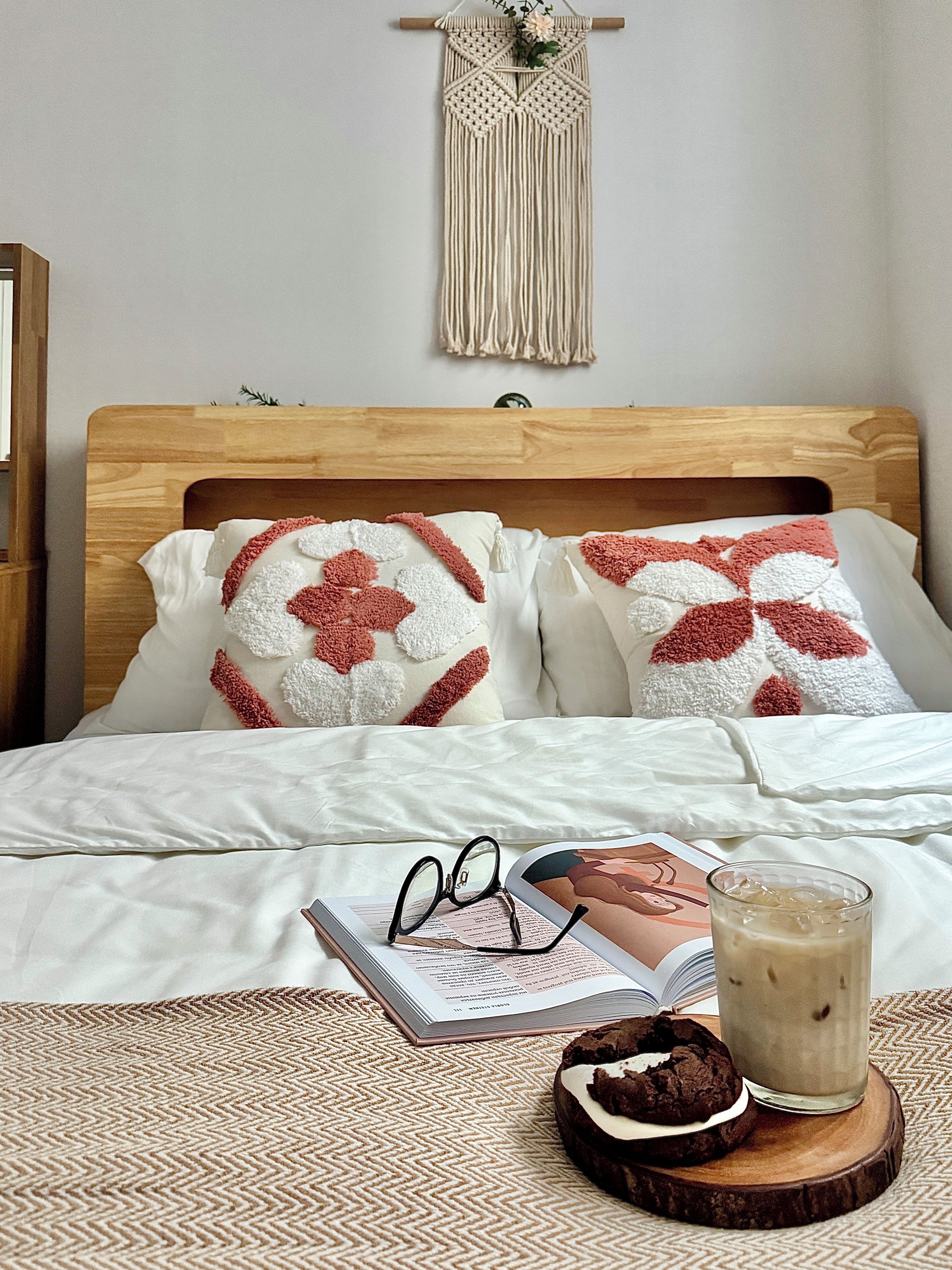 A neatly made bed with a wooden headboard, two decorative pillows, a book, a pair of glasses, and a glass of iced coffee on a coaster.
